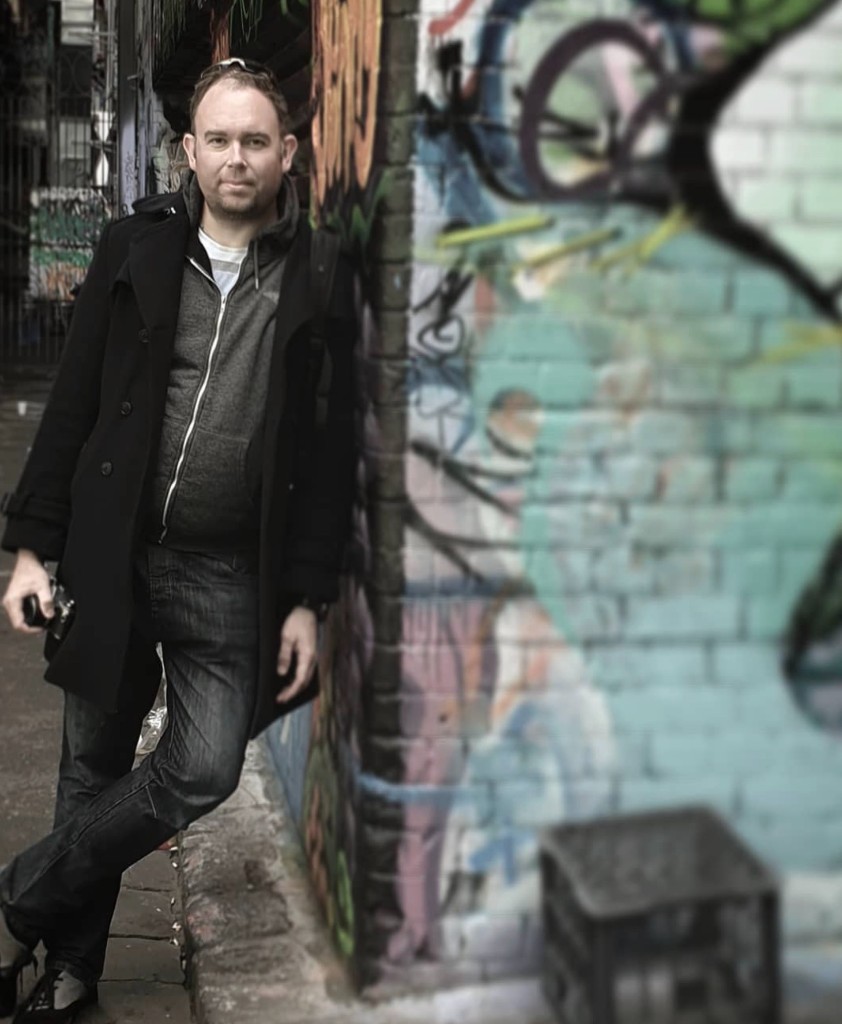 James Ian Gray in an alley beside graffiti-covered brickwork, holding a camera